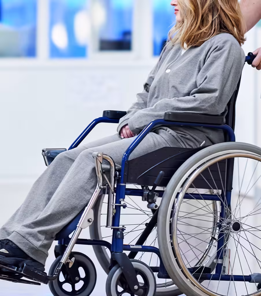 Manlig sjuksköterska kör en patient med patientkläder sittandes i rullstol i en sjukhuskorridor.